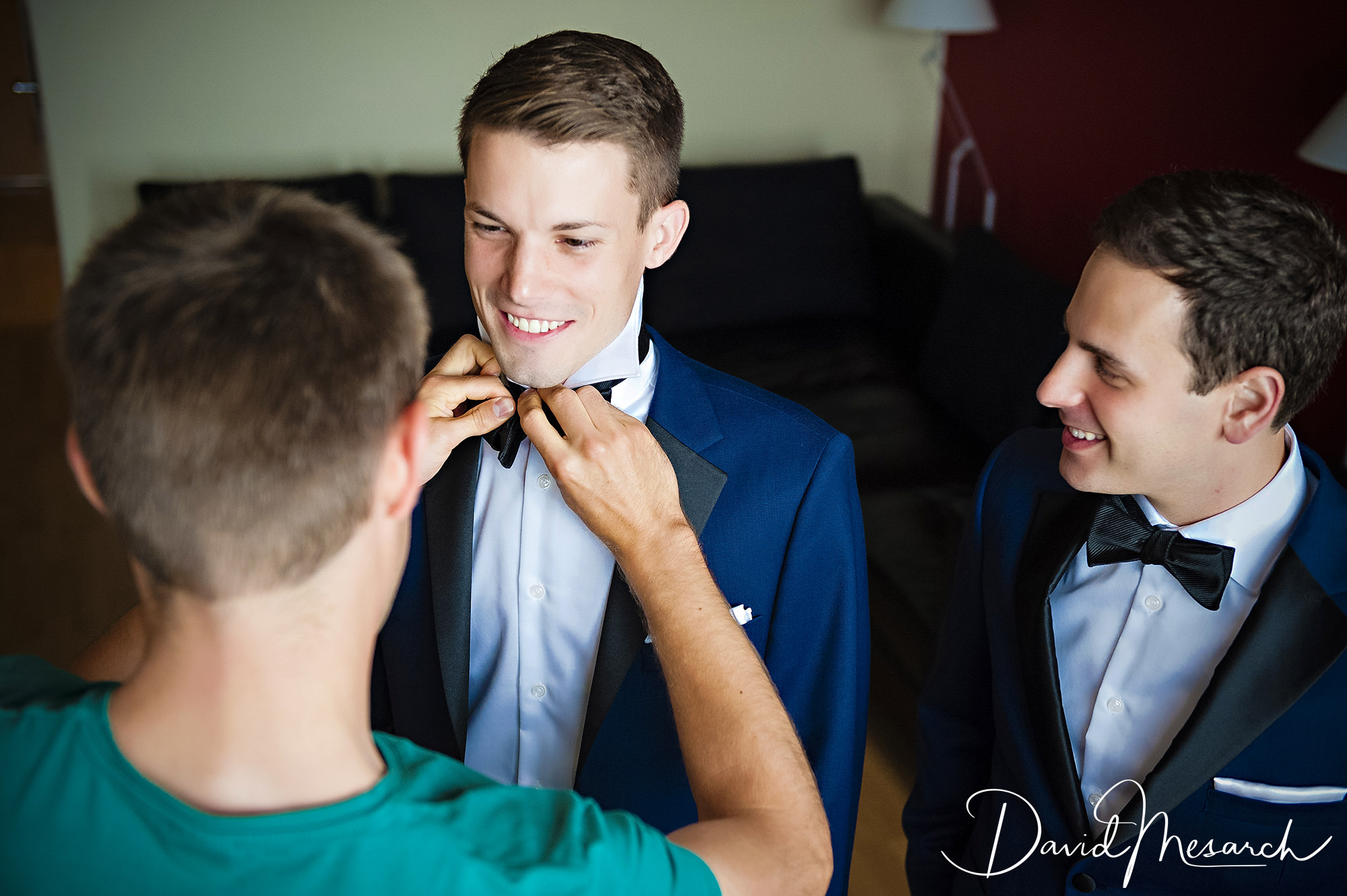 A groom smiles as his bow tie is adjusted while his partner watches during wedding preparations.