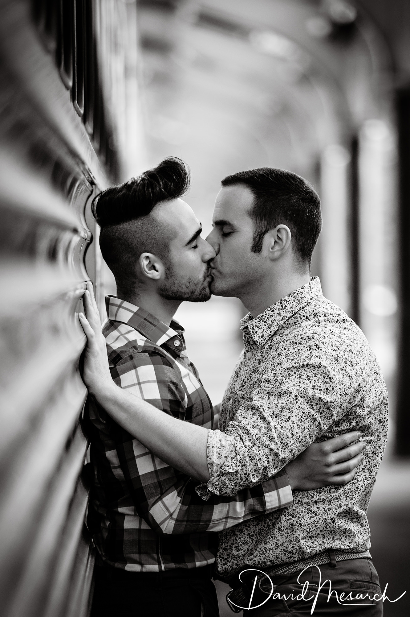 Two men kiss during a black-and-white portrait beside a reflective architectural wall.