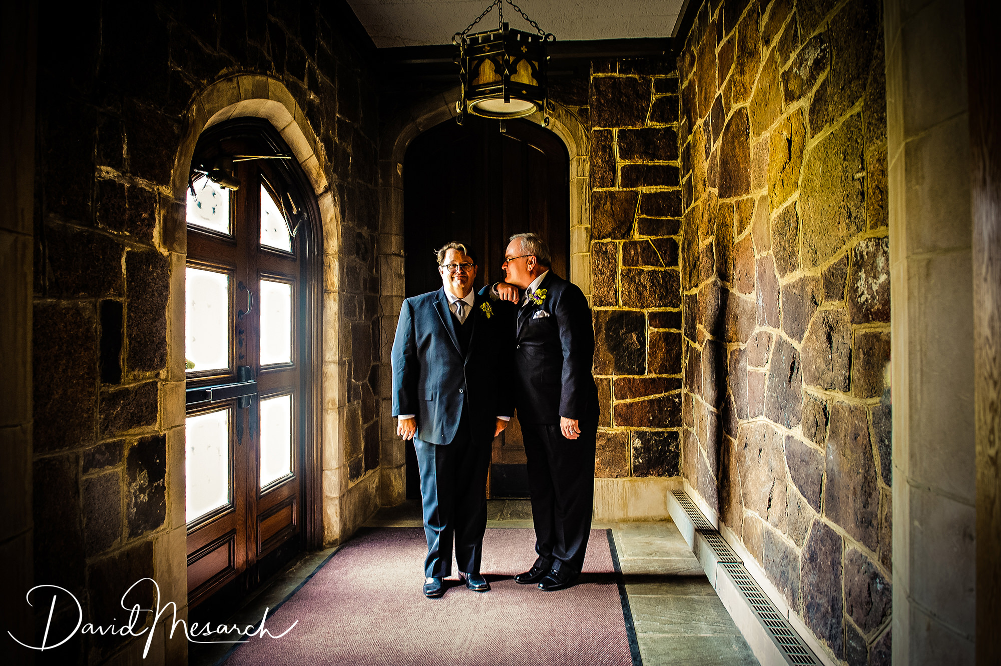 Two grooms stand together in a dramatic stone corridor, one leaning affectionately toward the other.