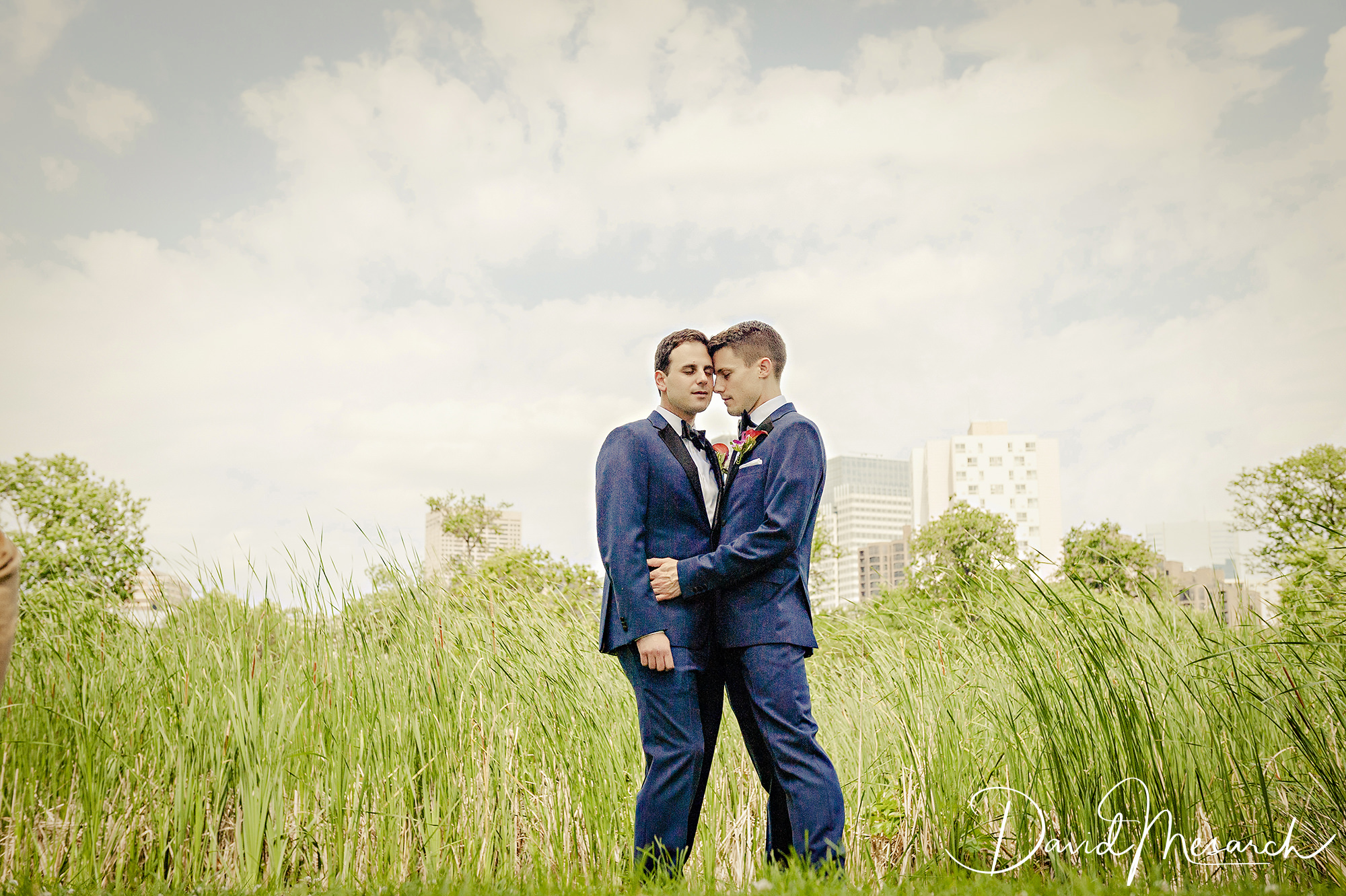 Two grooms in blue suits embrace in tall grass with the city skyline behind them.