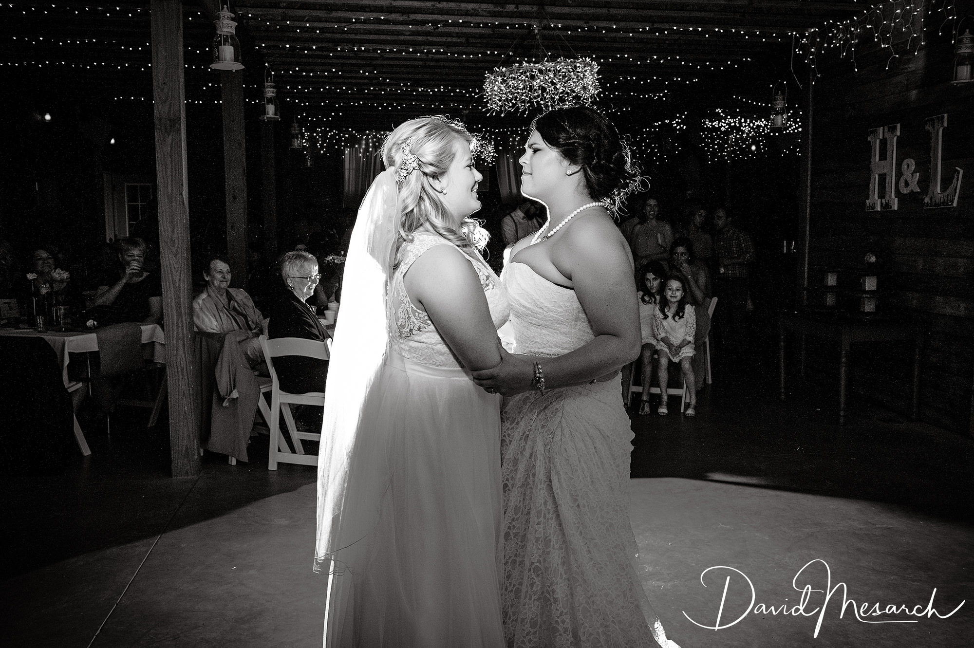 Two brides hold each other during their first dance beneath string lights while wedding guests look on.