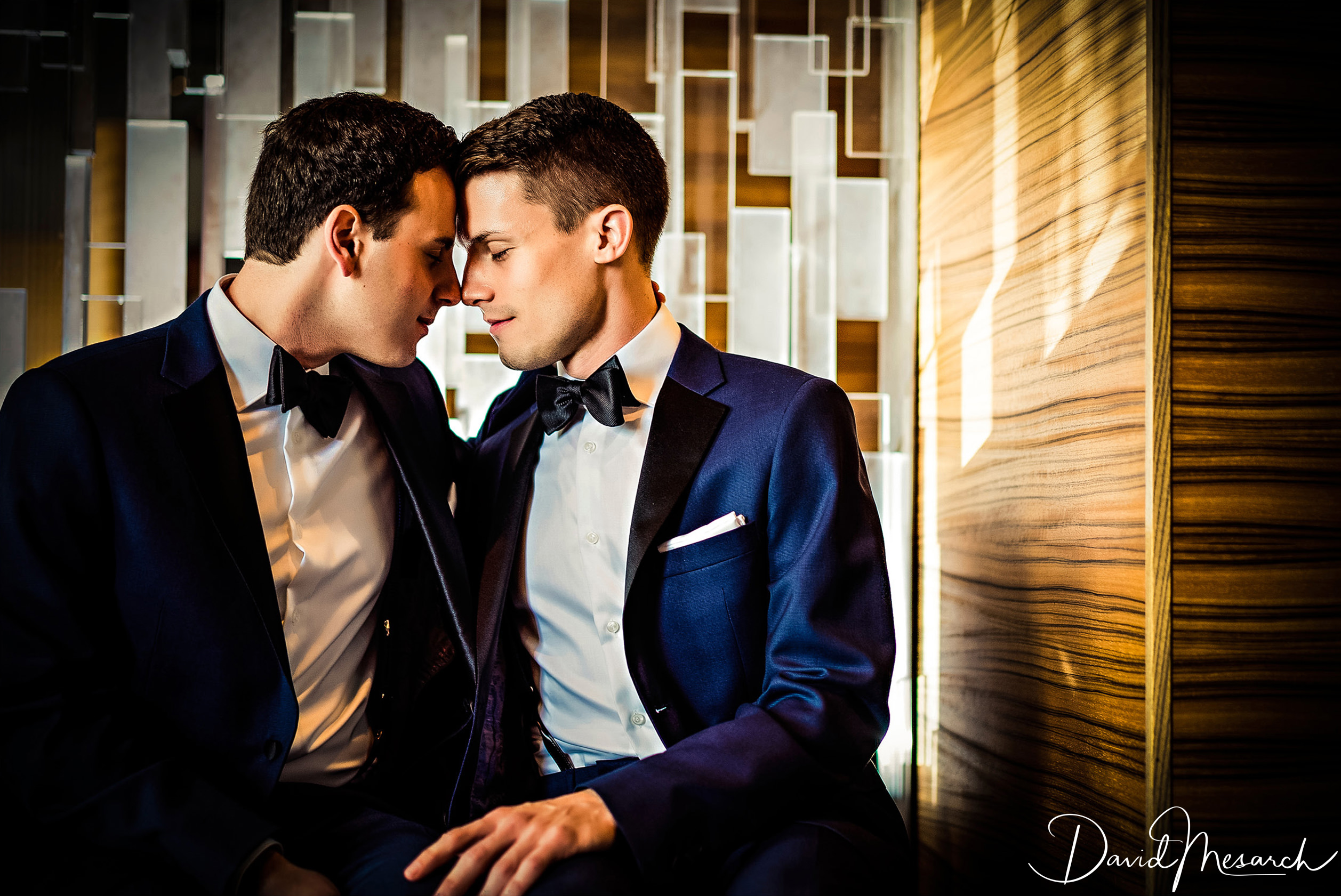 Two grooms in blue tuxedos sit close together during an editorial wedding portrait beside a geometric illuminated wall.